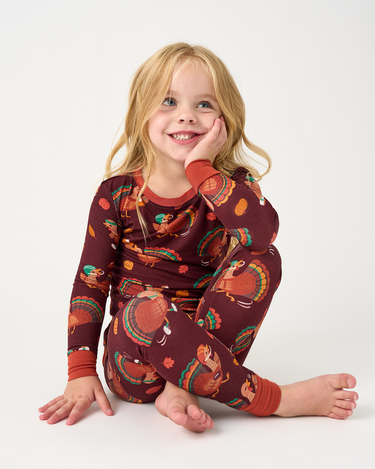 A young girl with blonde hair sits cross-legged on a white studio floor, wearing a long-sleeve pajama set with a dark red background and a colorful turkey print.