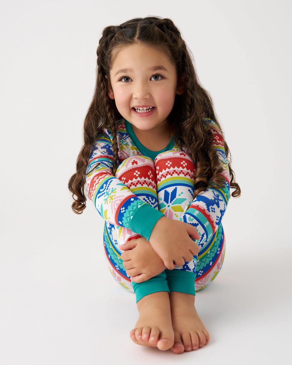 A young girl with long brown hair in braids sits cross-legged on a light gray studio background, wearing a colorful long-sleeve pajama set with a multicolor Fair Isle pattern and teal cuffs and collar.