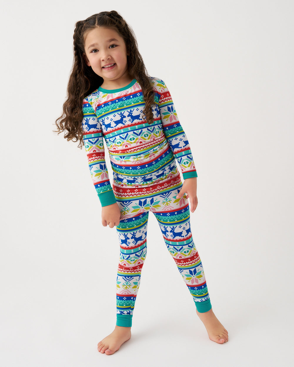 A young girl with long brown hair stands barefoot against a plain white studio background, wearing a long-sleeve matching pajama set with a colorful Nordic-style pattern in blue, red, green, yellow, and white.
