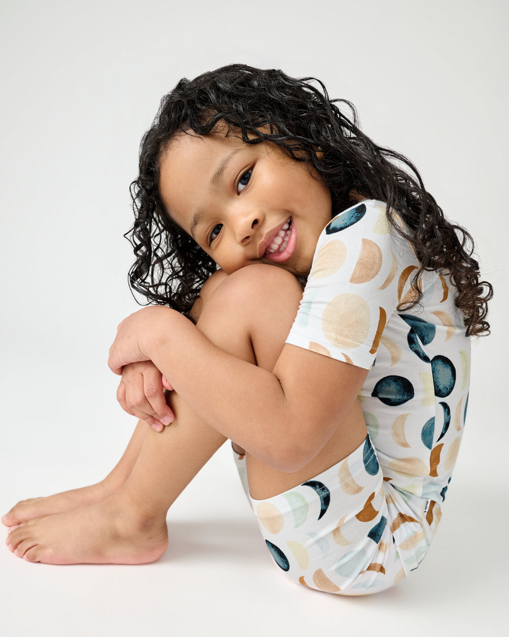 Sitting child holding her knees wearing the Luna Neutral Two-Piece Short Sleeve & Shorts Pajama Set