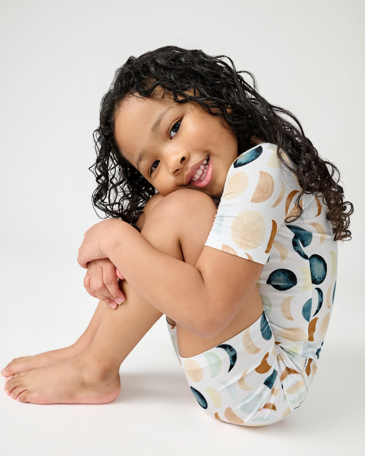 Sitting child holding her knees wearing the Luna Neutral Two-Piece Short Sleeve & Shorts Pajama Set