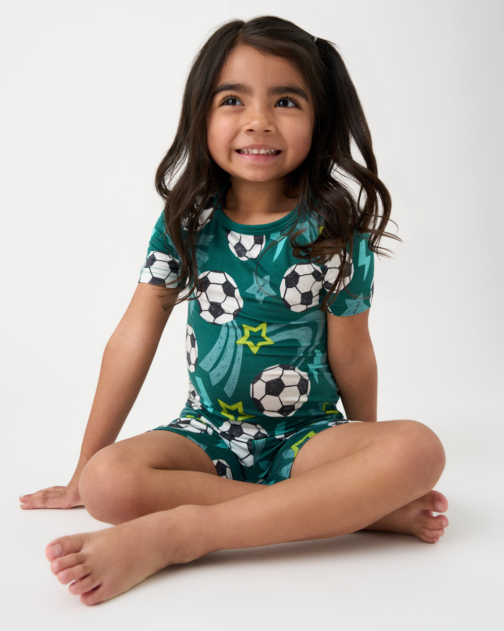 Sitting girl wearing the Green Soccer Stars Two-Piece Short Sleeve & Shorts Pajama Set