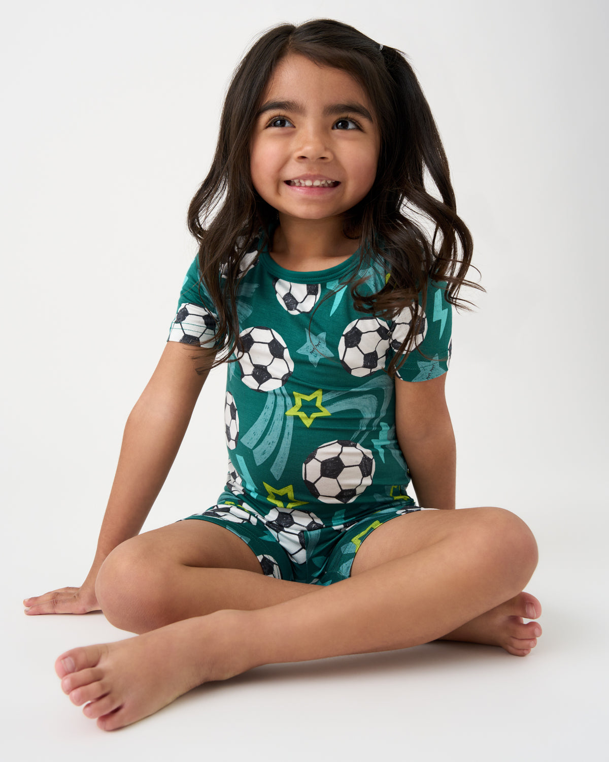 Sitting girl wearing the Green Soccer Stars Two-Piece Short Sleeve & Shorts Pajama Set