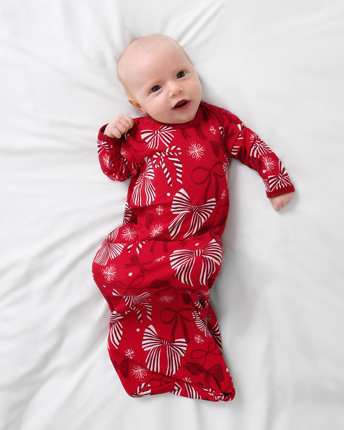 A baby lying on white bedding, wearing a red long-sleeve infant gown with a white and red bow pattern