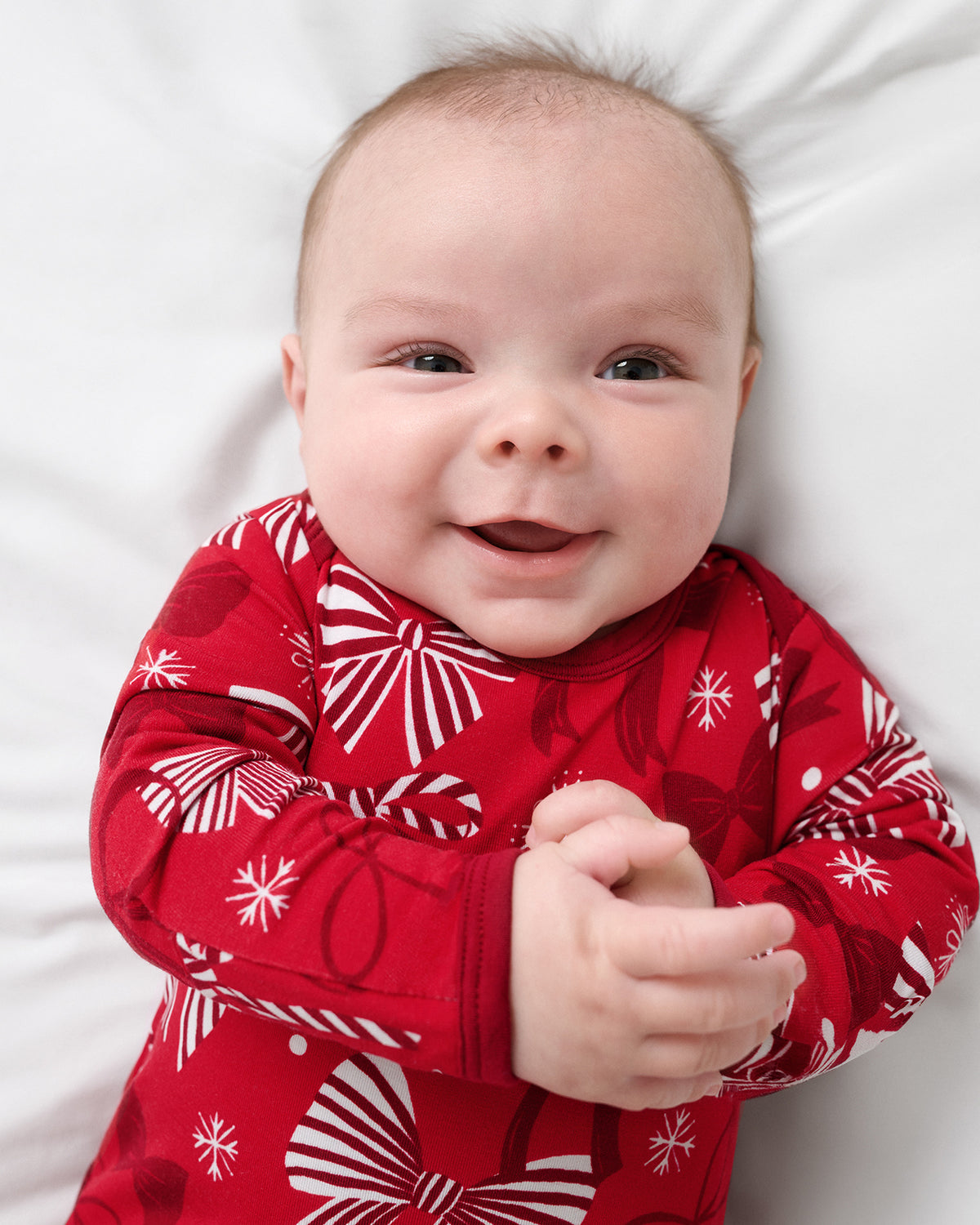 A smiling baby lying on a white pillow, wearing a red long-sleeve pajama top with a white holiday pattern of bows and snowflakes.