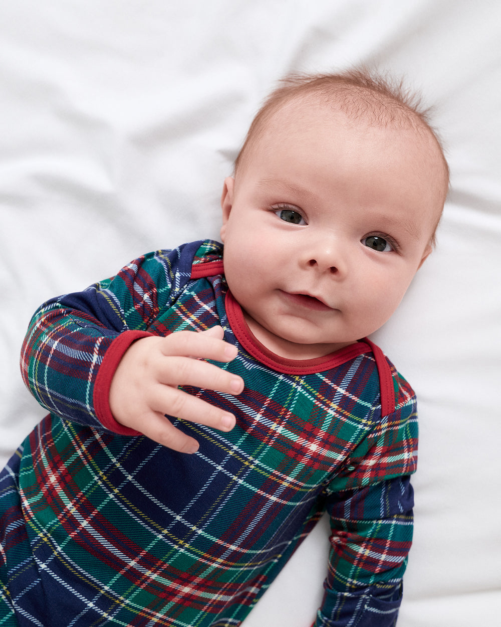 A baby lying on white bedding, wearing a long-sleeve plaid sleep gown in navy blue with green, red, and white tartan pattern and red trim.