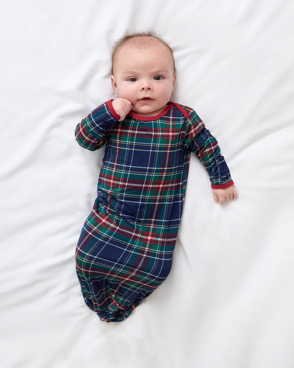 A baby lying on a white bed, wearing a long-sleeve sleep gown with a navy blue, green, and red plaid pattern.