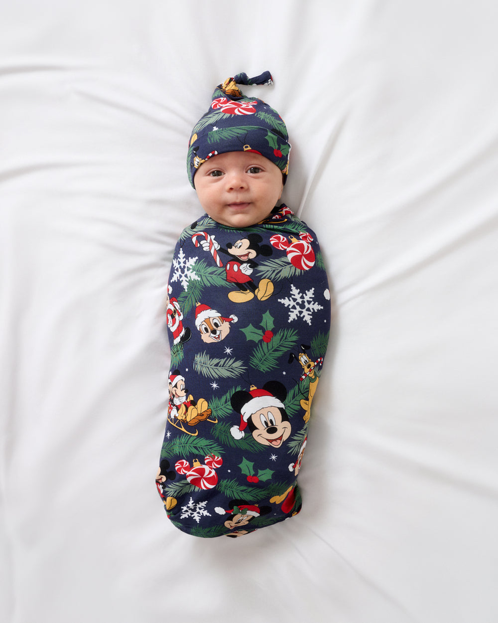 A baby swaddled in a navy blue holiday-themed blanket with Mickey Mouse and other Disney characters, lying on a white sheet. The baby is also wearing a matching navy blue hat with a knot top.