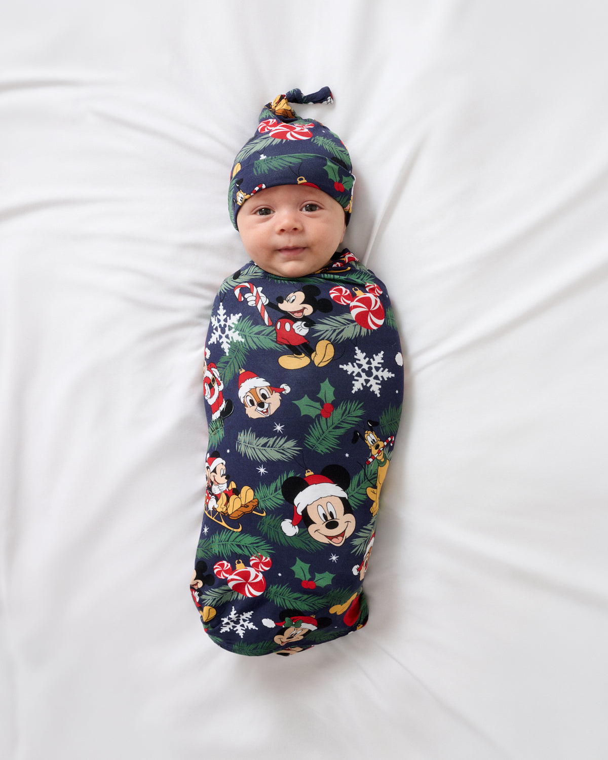 A baby swaddled in a navy blue holiday-themed blanket with Mickey Mouse and other Disney characters, lying on a white sheet. The baby is also wearing a matching navy blue hat with a knot top.