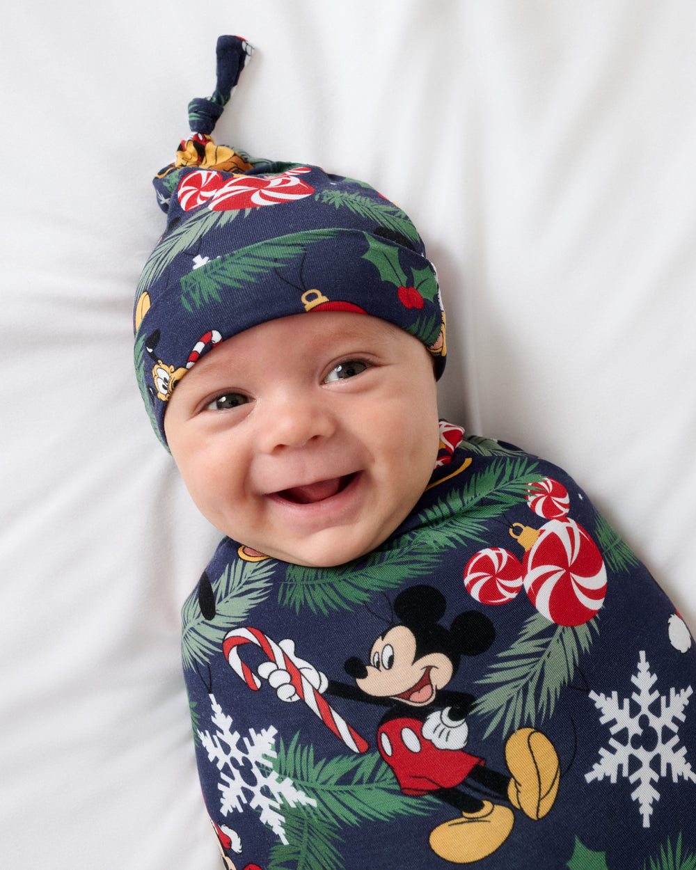 A smiling baby lying on a white blanket, wearing a navy blue Christmas-themed knit hat and matching swaddle blanket with Mickey Mouse, candy canes, pine branches, holly, and snowflakes.
