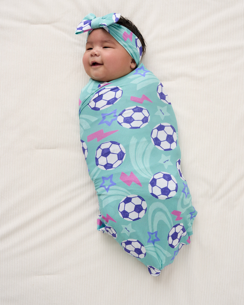 Aerial view of newborn wearing the Aqua Soccer Stars Swaddle & Luxe Bow Headband Set