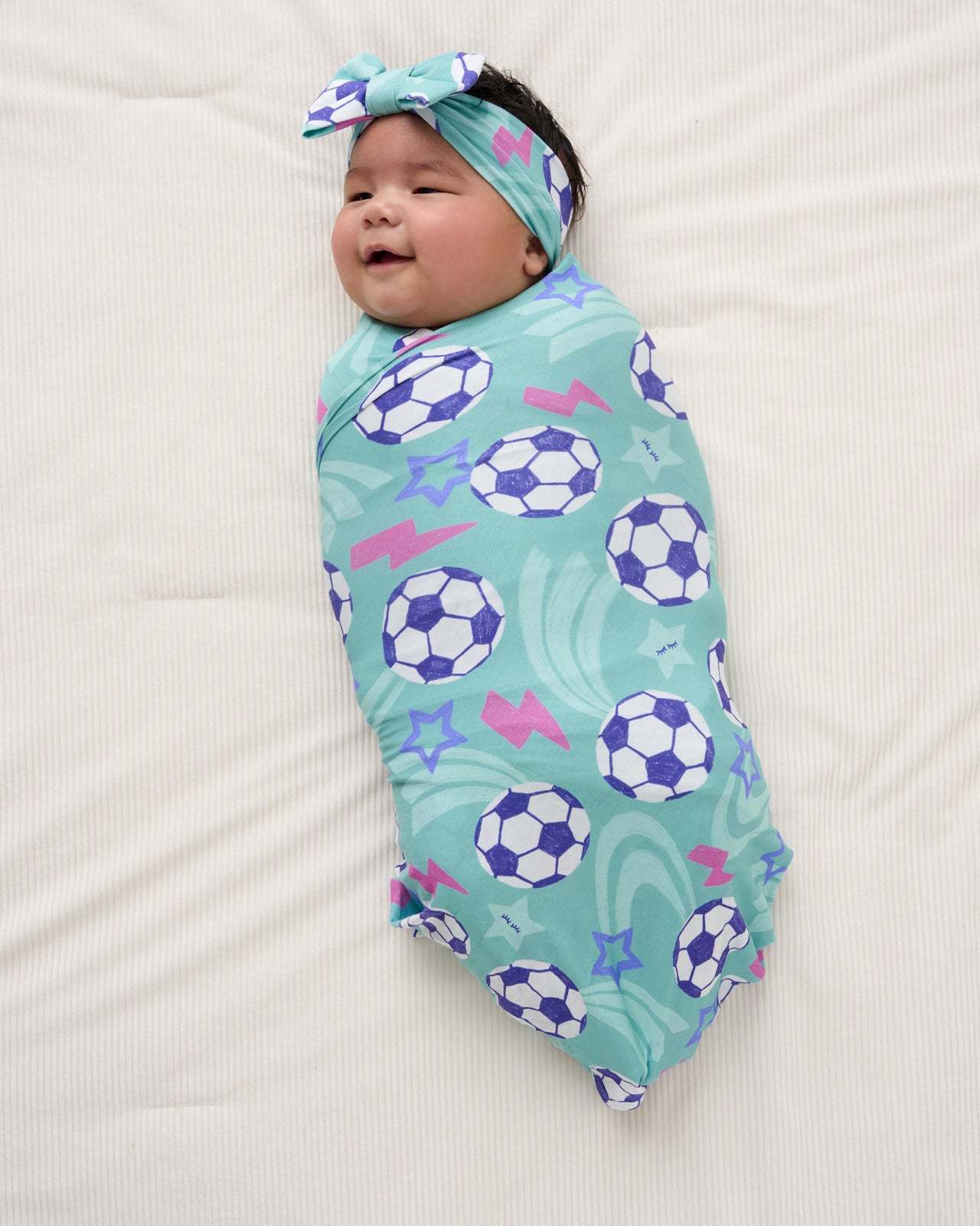 Aerial view of newborn wearing the Aqua Soccer Stars Swaddle & Luxe Bow Headband Set