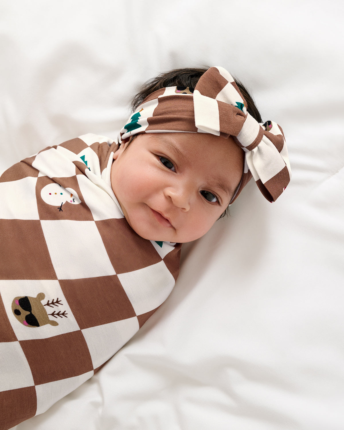 A baby lying on white bedding, swaddled in a brown and cream checkered wrap blanket with small holiday-themed prints, and wearing a matching checkered bow headband.
