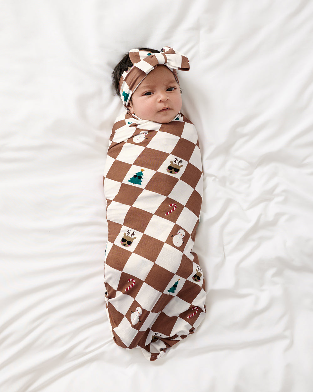A baby swaddled in a brown and cream checkered blanket with small holiday motifs, wrapped in a matching headband, lying on a white bed.