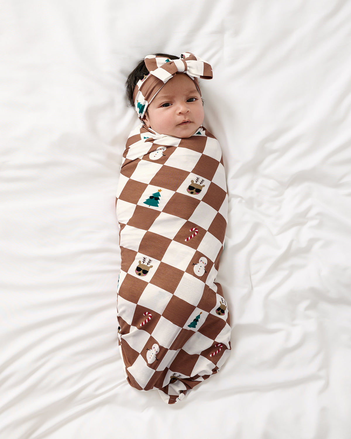 A baby swaddled in a brown and cream checkered blanket with small holiday motifs, wrapped in a matching headband, lying on a white bed.