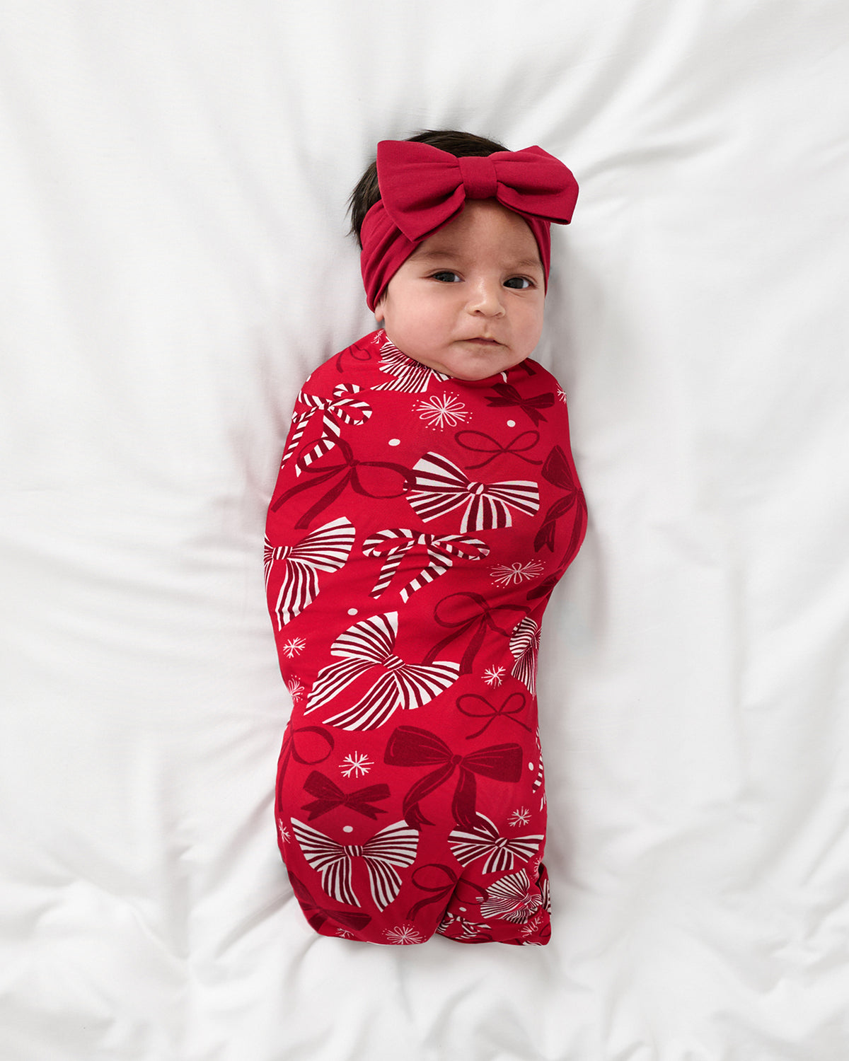 A baby wrapped in a red holiday-themed swaddle blanket with white and red bow patterns, wearing a matching red bow headband, lying on a white fabric background.