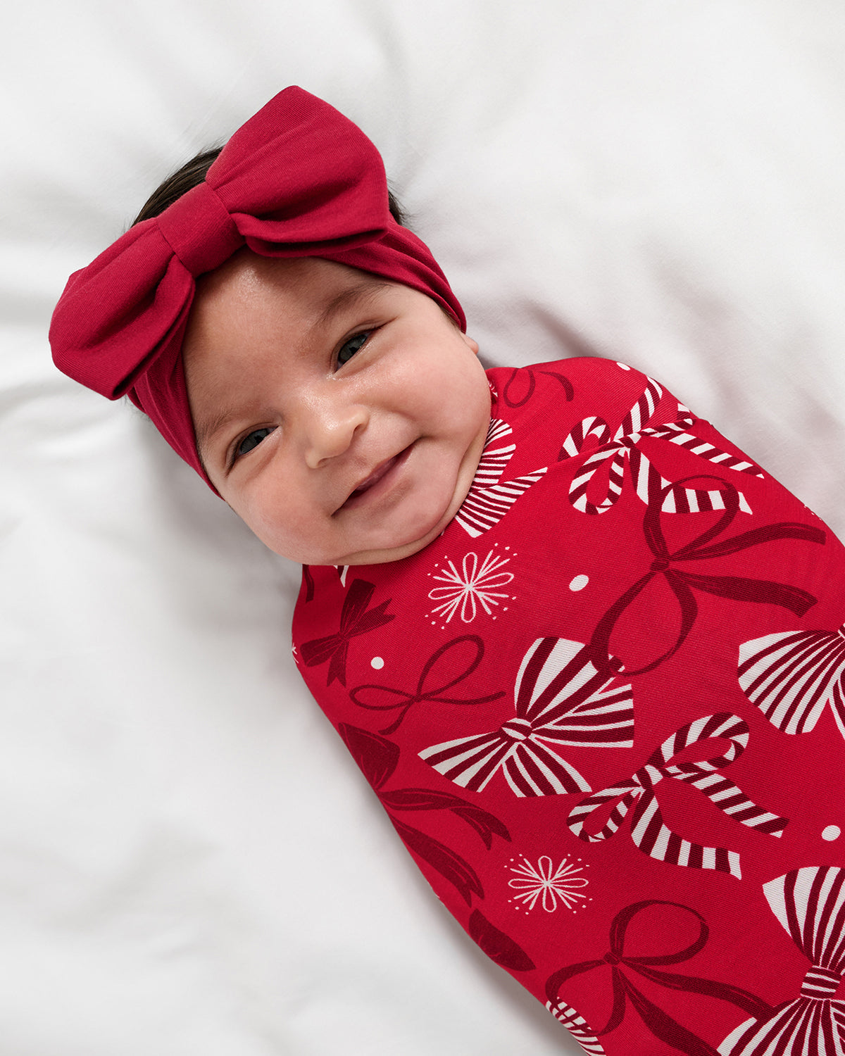 A baby lying on a white blanket, wearing a large red bow headband and a red swaddle blanket with a white and red bow pattern.