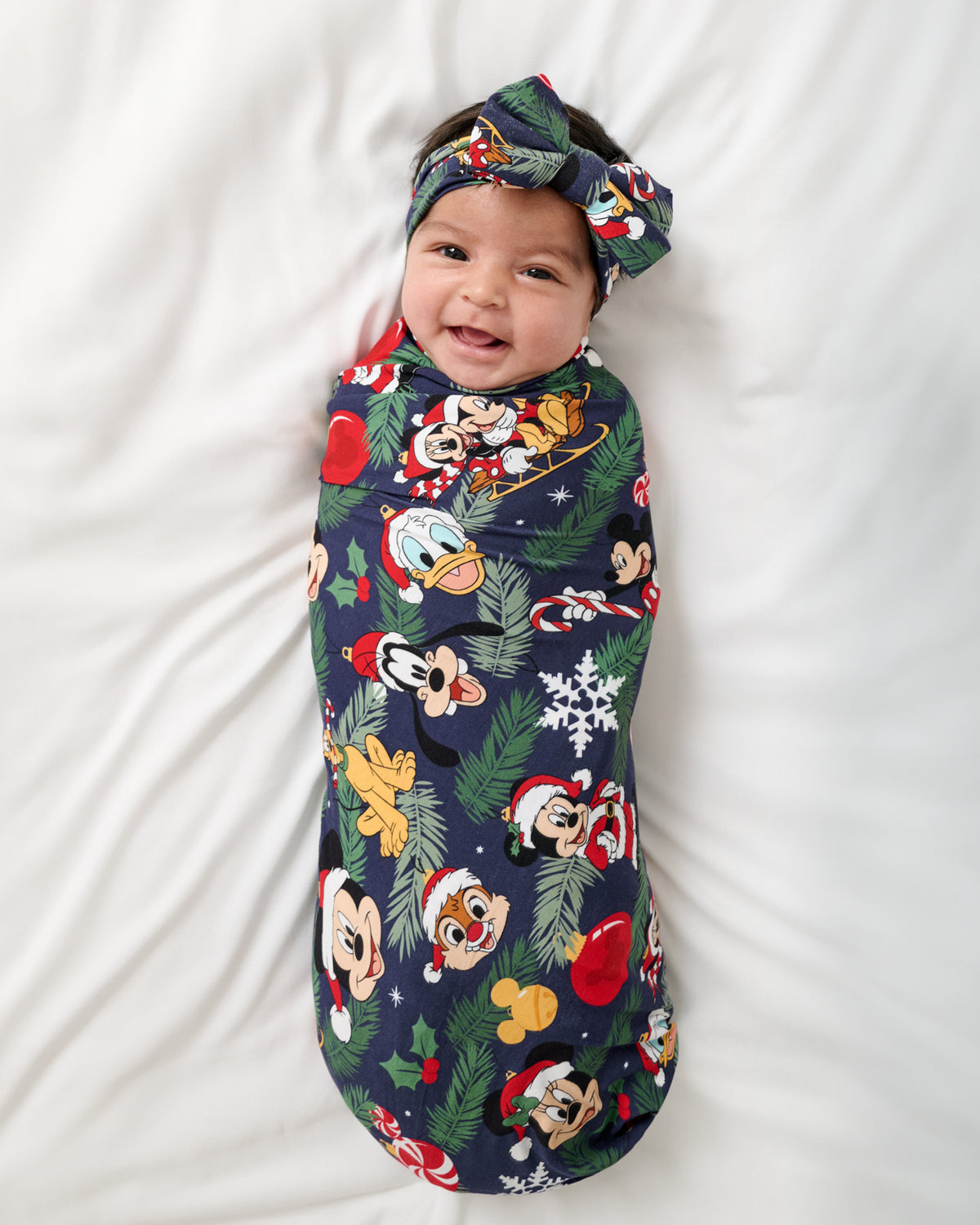 A baby wrapped in a navy blue Christmas-themed swaddle blanket, wearing a matching knotted headband, lying on a white fabric background.