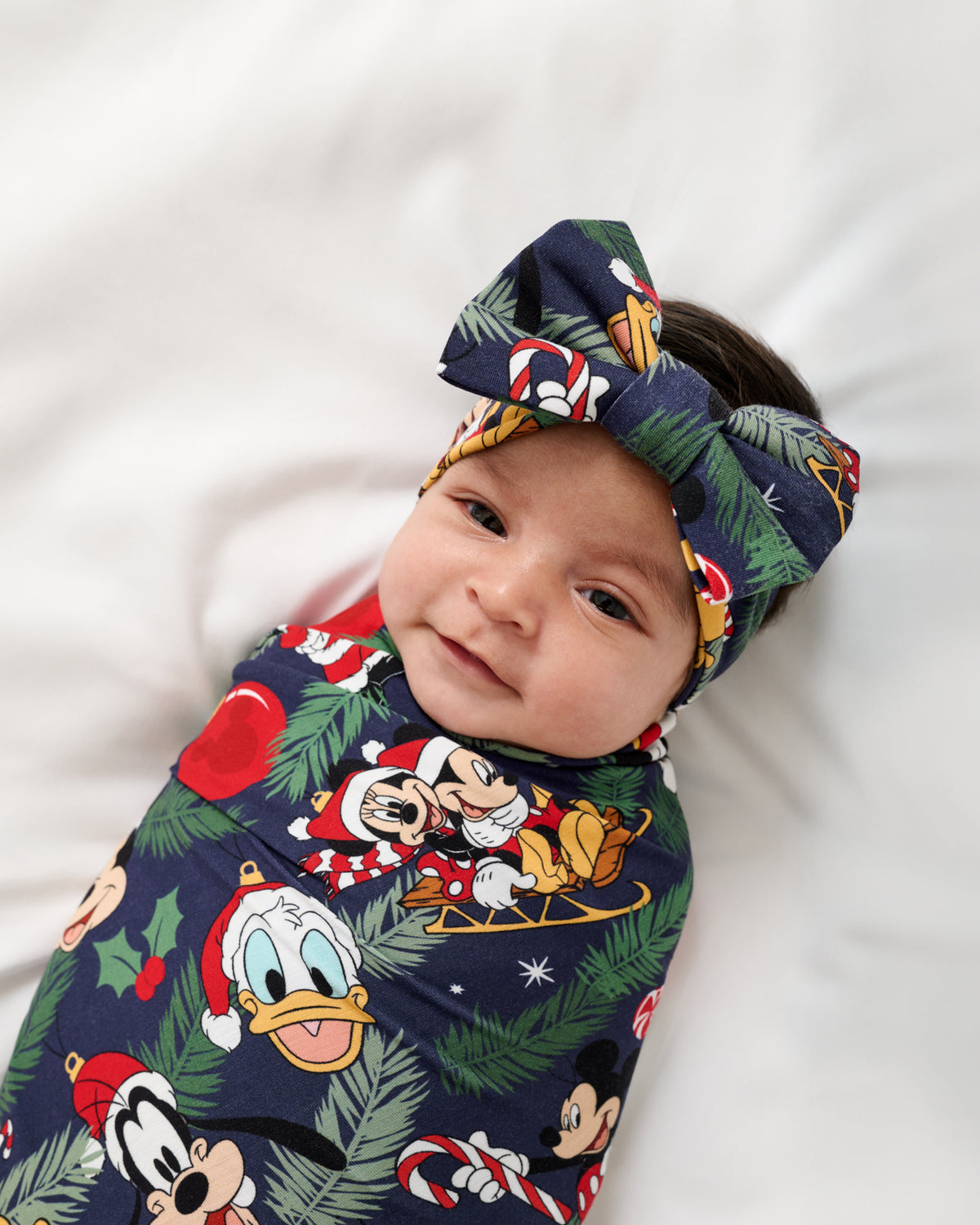 A baby swaddled in a navy blue Disney holiday print wrap with a matching bow headband, lying on a white background.