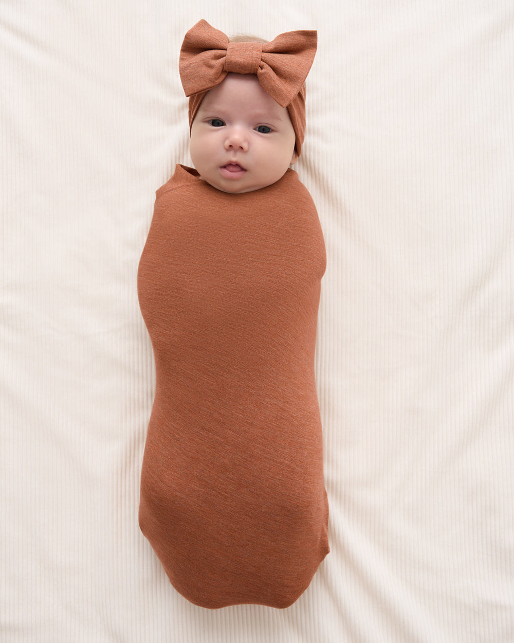 A baby swaddled in a rust-brown fabric wrap and wearing a matching brown bow headband, lying on a cream-colored ribbed blanket.