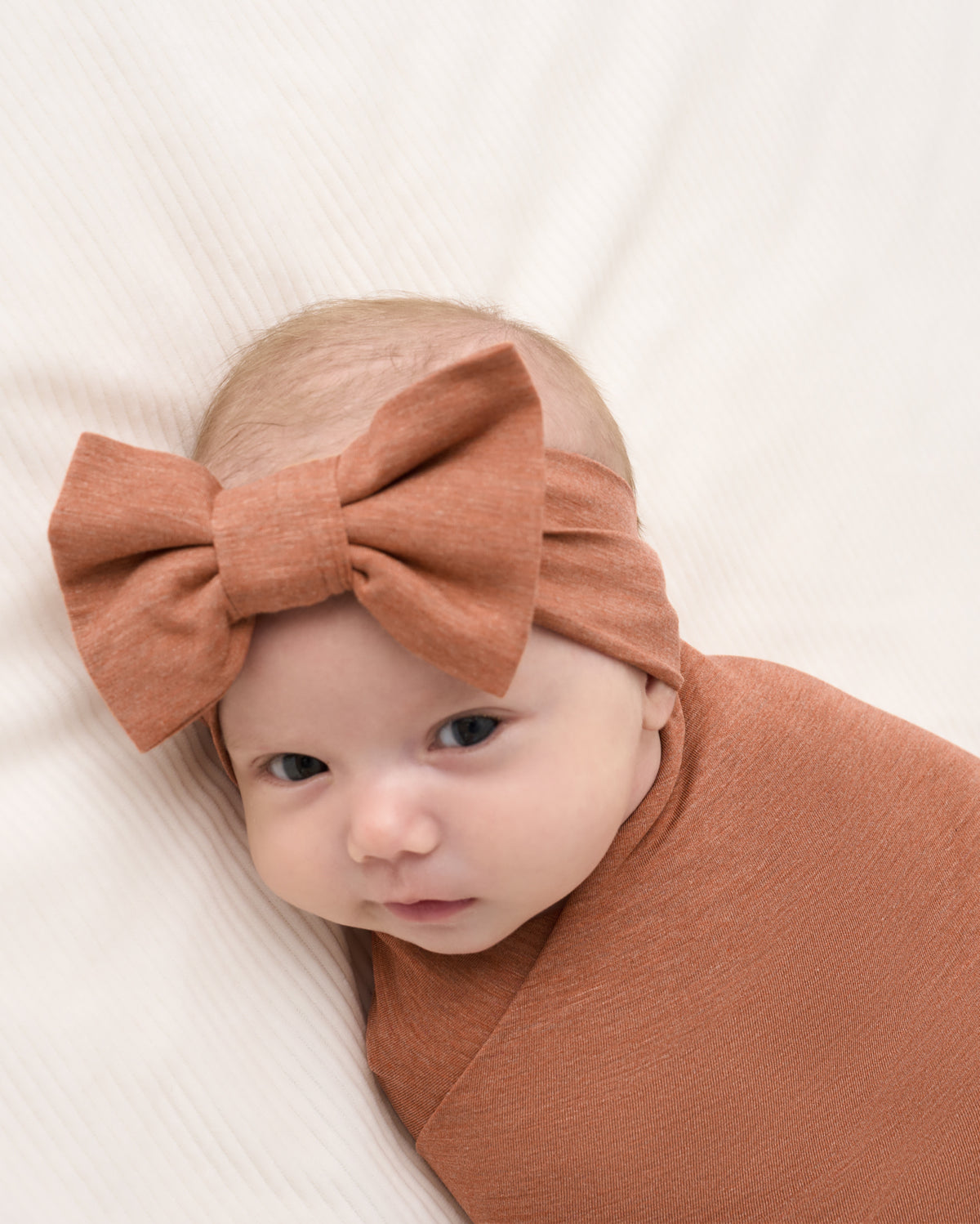 A baby swaddled in a rust-colored blanket and wearing a matching large bow headband, lying on a textured cream blanket.