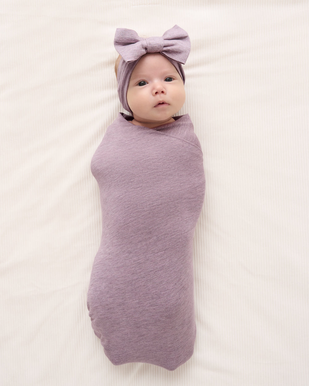 A baby swaddled in a dusty purple blanket and wearing a matching purple bow headband, lying on a white textured blanket.