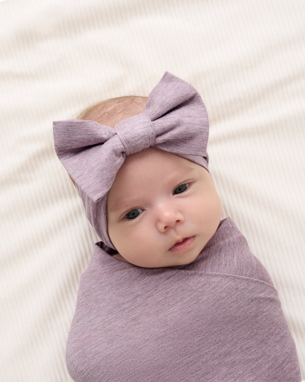 A baby swaddled in a matching dusty purple wrap and wearing a matching bow headband, lying on a textured cream blanket.