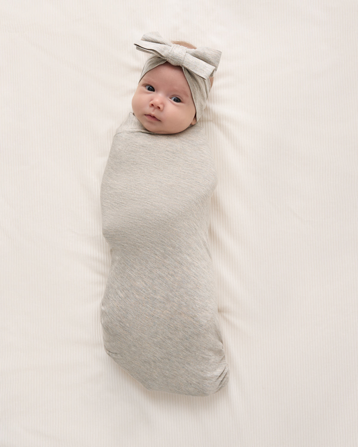 A baby wrapped in a light gray heather swaddle blanket, wearing a matching gray bow headband, lying on a white textured blanket.