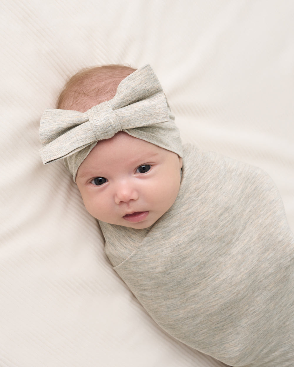 A baby swaddled in a light gray heathered swaddle blanket and wearing a matching light gray bow headband, lying on a white textured blanket.