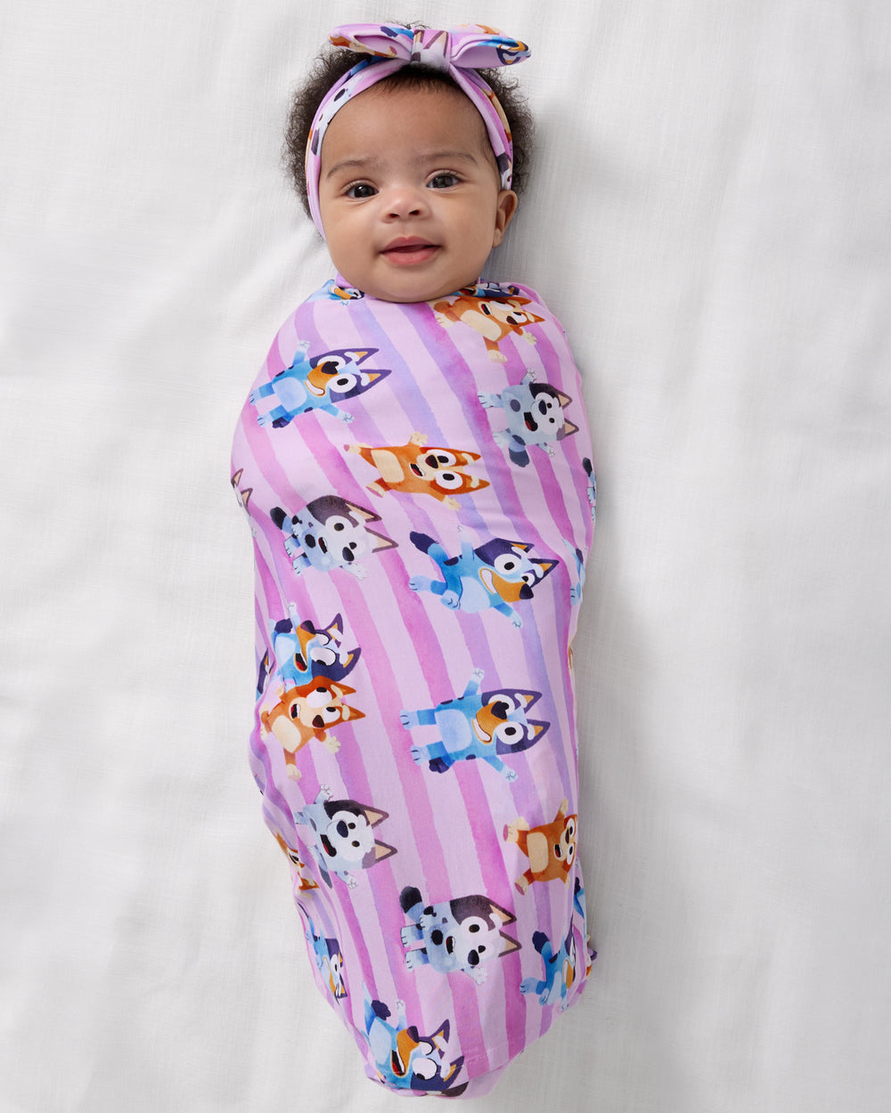Aerial view of baby wrapped in the Pink Bluey Dance All Day Swaddle & Luxe Bow Headband Set
