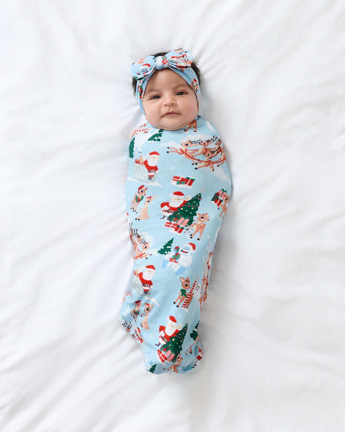 A baby swaddled in a light blue Christmas-themed blanket with Santa Claus and trees, wearing a matching headband, lying on a white bed.
