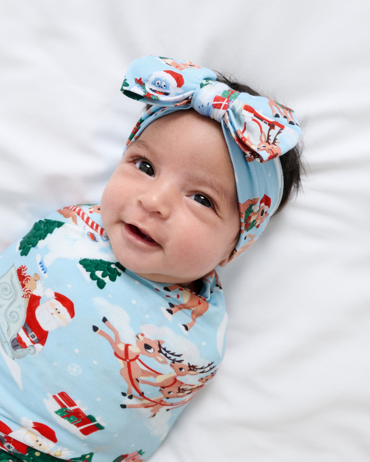 A baby lying on a white blanket, wearing a light blue Christmas-themed swaddle wrap and a matching bow headband with Santa Claus, reindeer, and snowflake patterns.