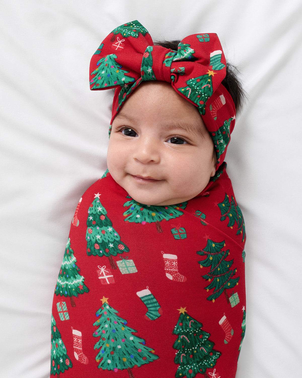 A baby wrapped in a festive red swaddle blanket with a green Christmas tree and stocking pattern, wearing a matching red bow headband, lying on a white background.