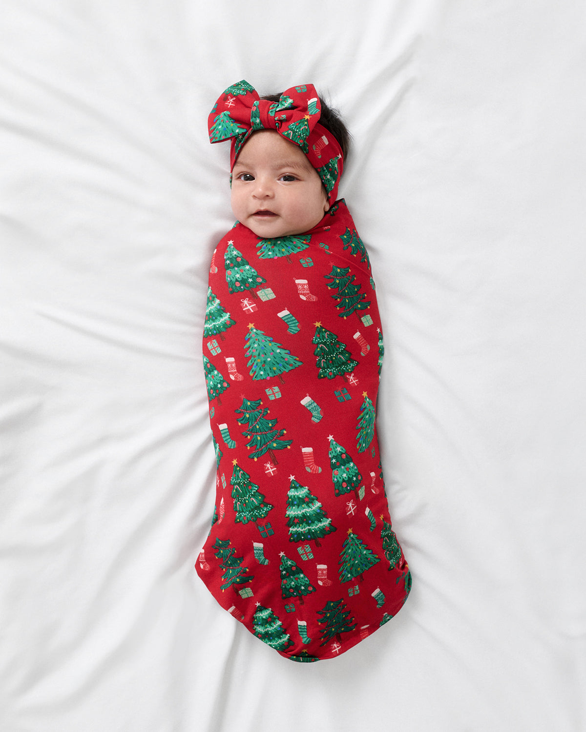 A baby swaddled in a red Christmas-themed blanket with green trees and small gift prints, wearing a matching red bow headband, lying on a white sheet.