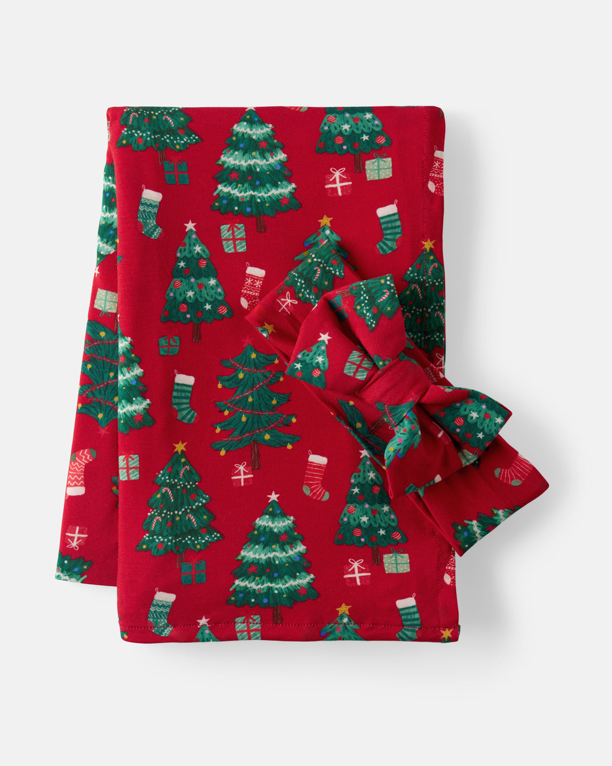 A folded red fabric item, likely a blanket or towel, featuring a festive Christmas print with green trees, stockings, and gifts, tied with a matching red and green bow on a light gray background.