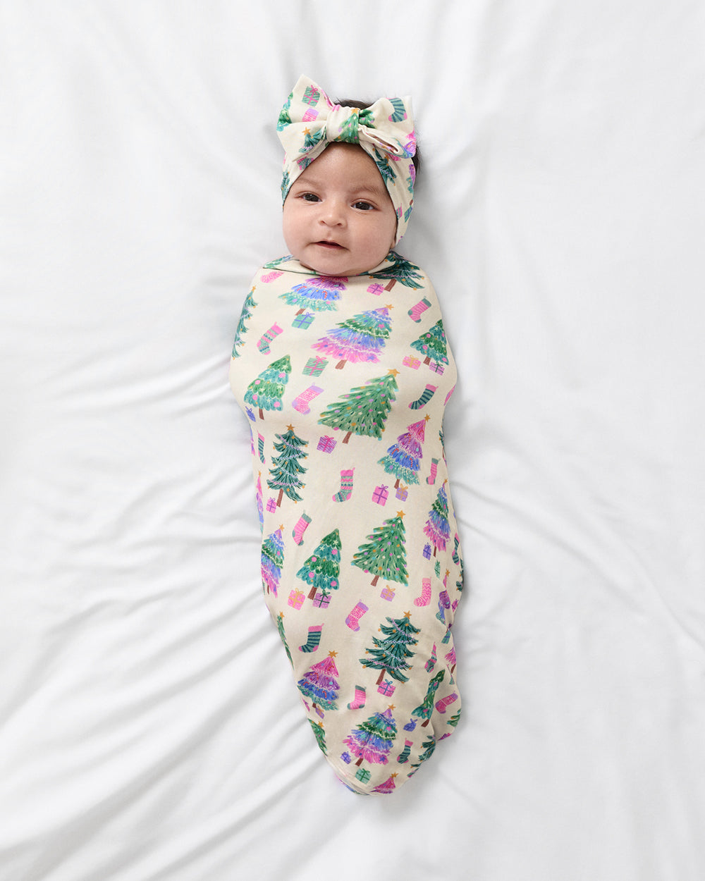 A baby swaddled in a cream-colored blanket with a green Christmas tree and pink gift pattern, wearing a matching green and pink bow headband, lying on a white sheet.