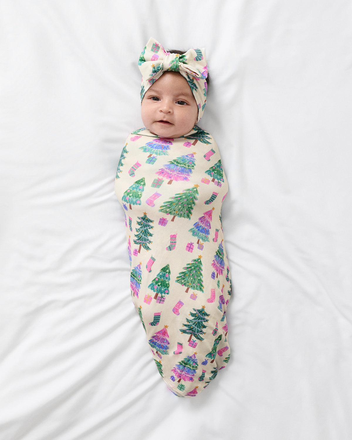 A baby swaddled in a cream-colored blanket with a green Christmas tree and pink gift pattern, wearing a matching green and pink bow headband, lying on a white sheet.
