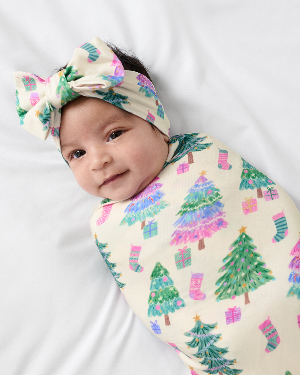 A baby swaddled in a cream-colored blanket with a colorful Christmas tree and stocking pattern, wearing a matching cream headband with a large bow, lying on a white background.