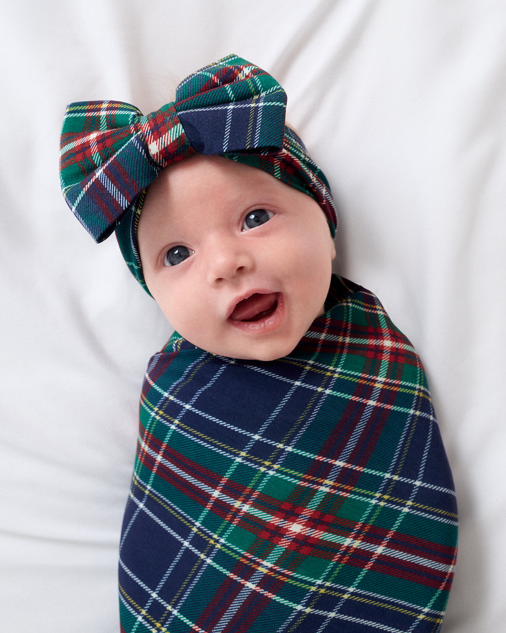 A baby swaddled in a green, navy, and red tartan plaid wrap, lying on a white background and wearing a matching plaid bow headband.