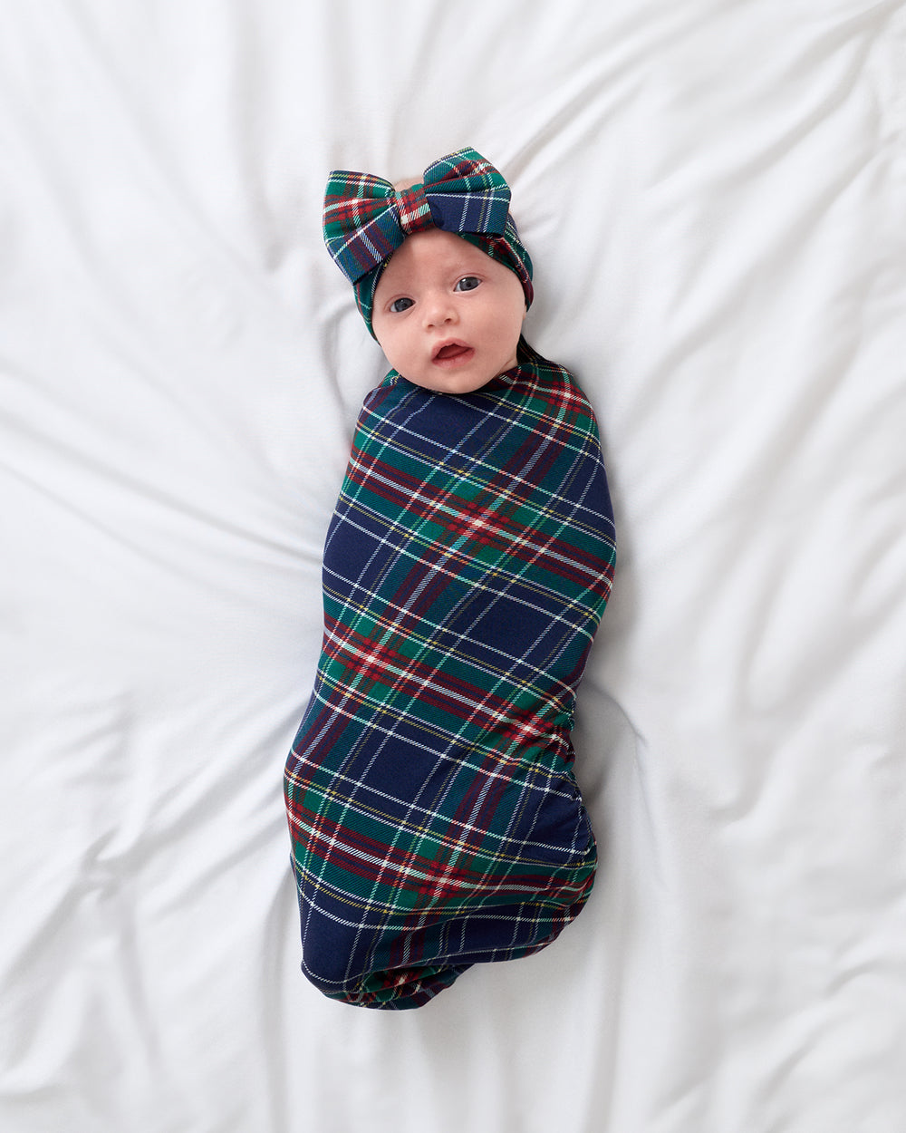 A baby swaddled in a navy blue tartan blanket with red, green, and white stripes, wearing a matching bow headband, lying on a white bed.