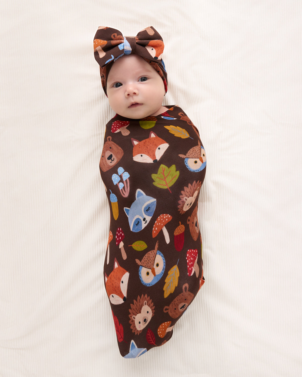 A baby swaddled in a brown fabric swaddle blanket with a colorful woodland print of foxes, owls, hedgehogs, and mushrooms, wearing a matching bow headband, lying on a white textured blanket.