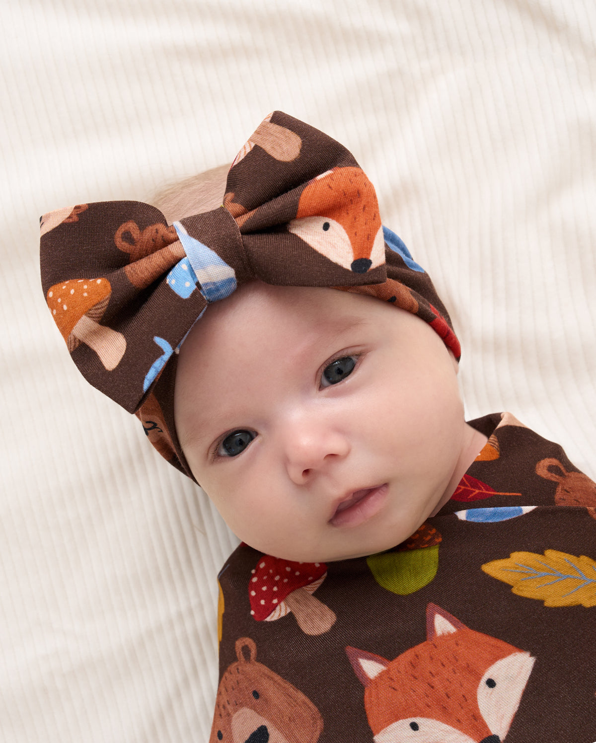 A baby lying on a cream-colored ribbed blanket, wearing a brown fabric headband and matching baby wrap with a colorful woodland print of foxes, mushrooms, and leaves.