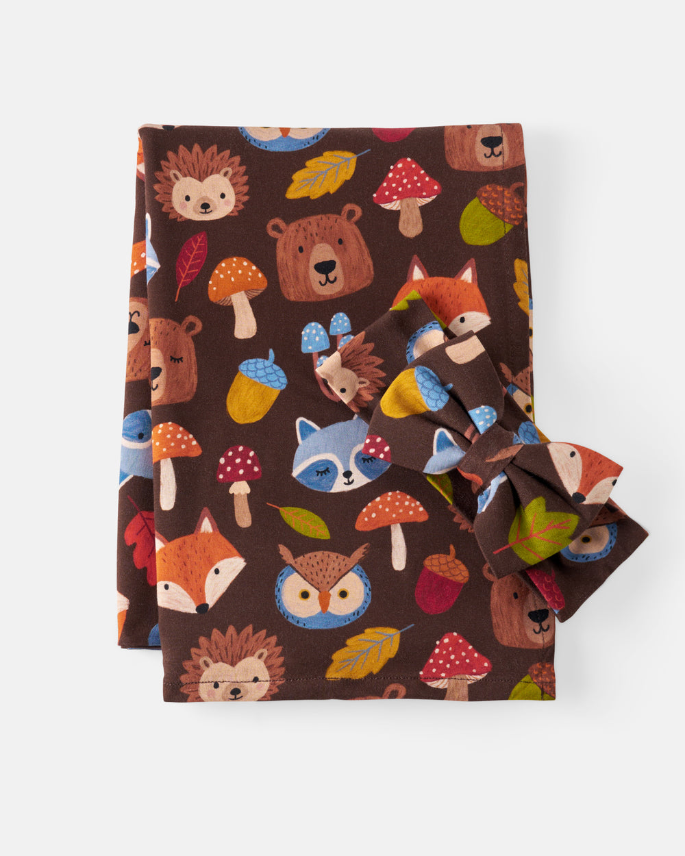 A folded blanket and bow set, featuring a woodland animal print on a dark brown background. The pattern includes foxes, hedgehogs, owls, bears, mushrooms, acorns, and leaves in orange, red, blue, yellow, and green.