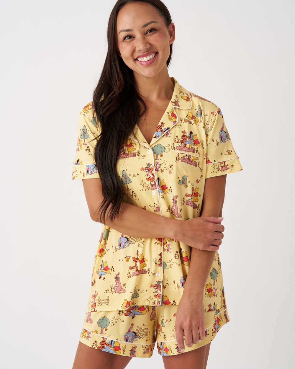 A woman modeling a two-piece pajama set featuring a short-sleeve button-up top and matching shorts in a pale yellow color with a whimsical multicolor cartoon print, set against a plain light gray background.