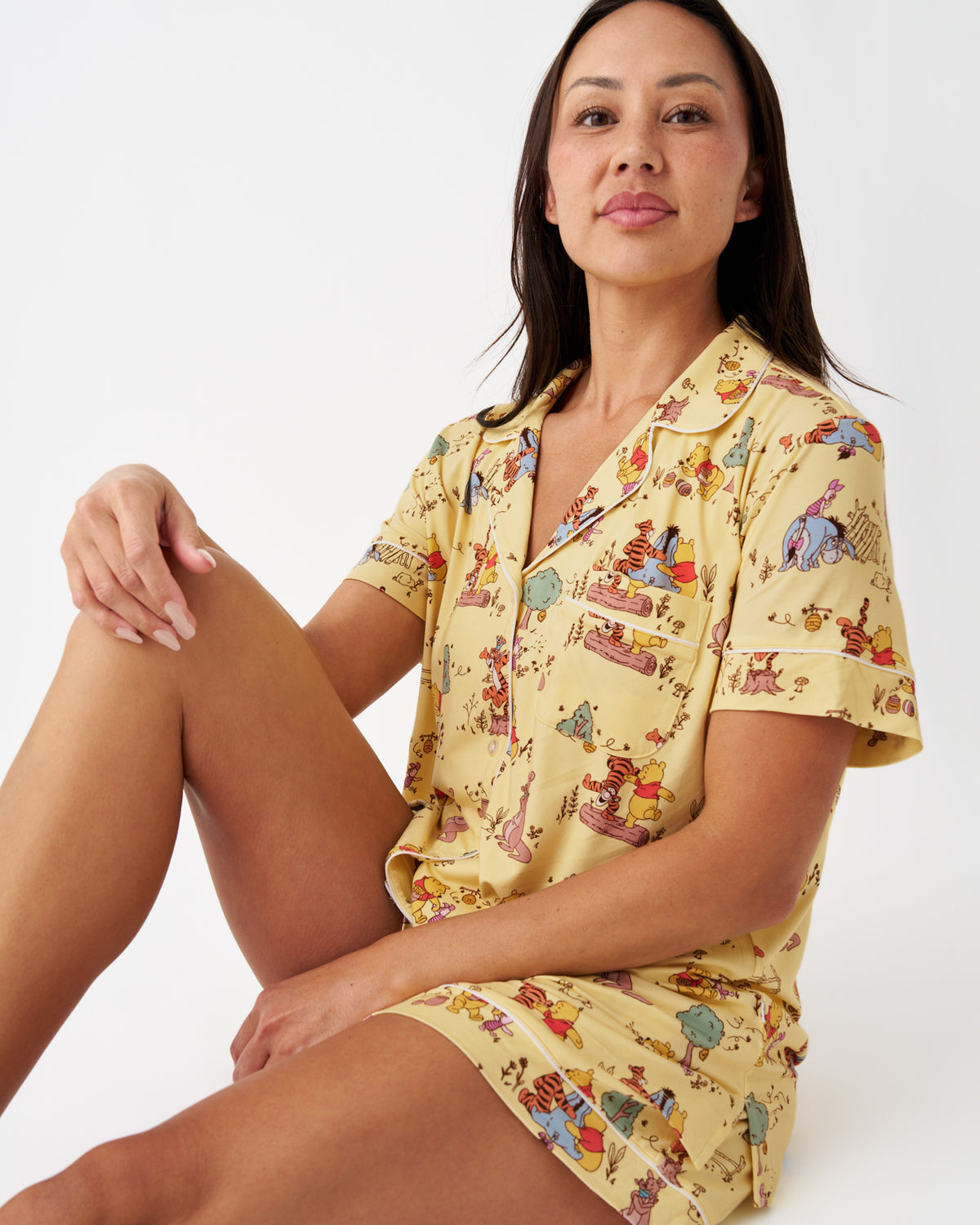 A woman wearing a short-sleeve pajama shirt and matching shorts set in a pale yellow color with a colorful cartoon character print, posed against a plain white studio background.