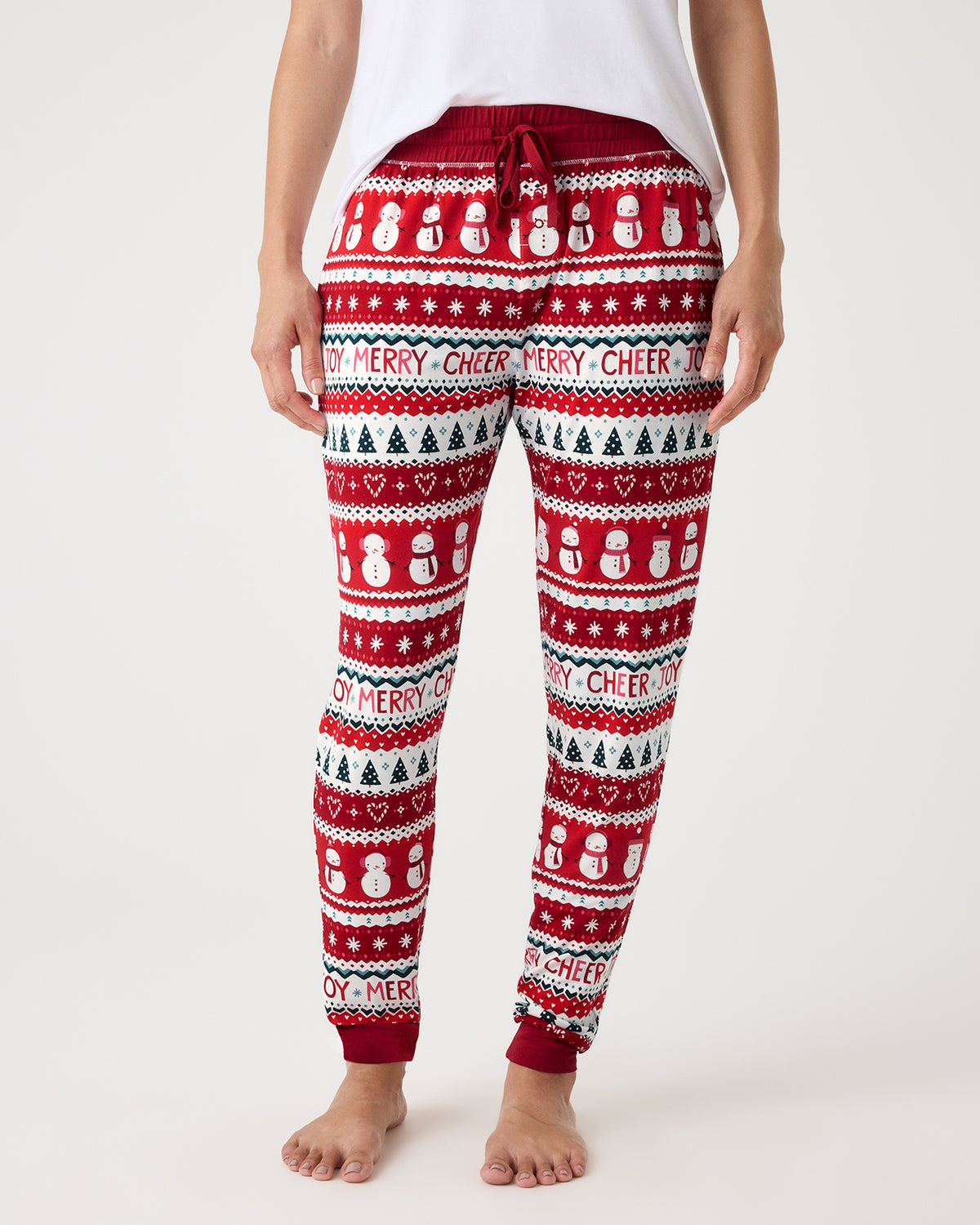 A person wearing red, white, and green holiday sweatpants with snowman and Christmas tree patterns, shown from the waist down against a light gray studio background.