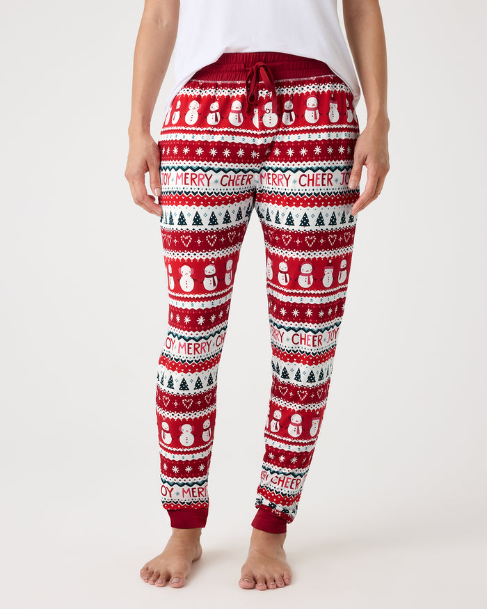 A person wearing red, white, and green holiday sweatpants with snowman and Christmas tree patterns, shown from the waist down against a light gray studio background.