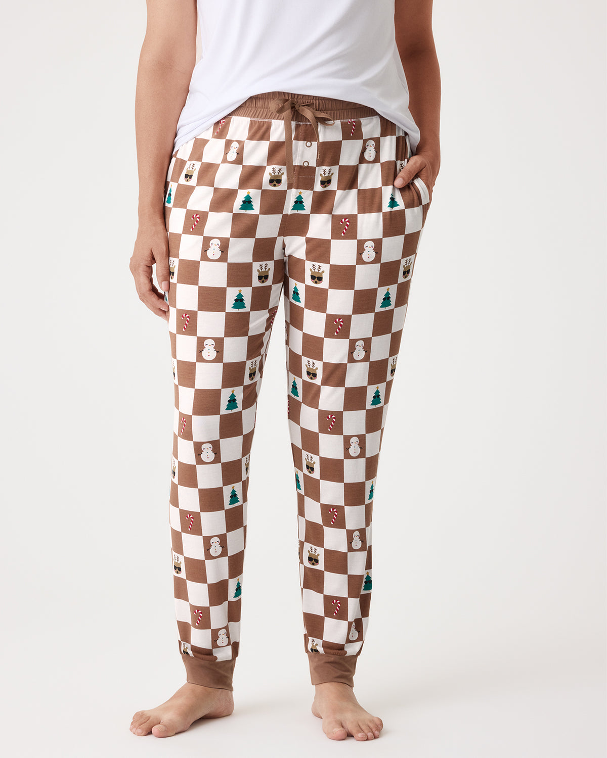 A person wearing brown and cream checkered pajama pants with small holiday-themed icons printed throughout, standing barefoot against a light gray studio background.