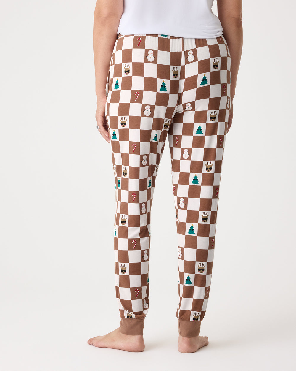 A person shown from the back wearing brown and cream checkered pajama pants with small holiday-themed icons printed on them, standing barefoot against a light gray studio background.