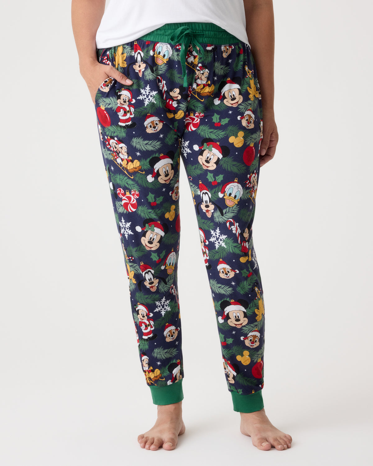A person wearing holiday-themed lounge pants with an all-over print of Mickey Mouse faces, Santa hats, snowflakes, and Christmas motifs, shown against a plain light gray studio background.